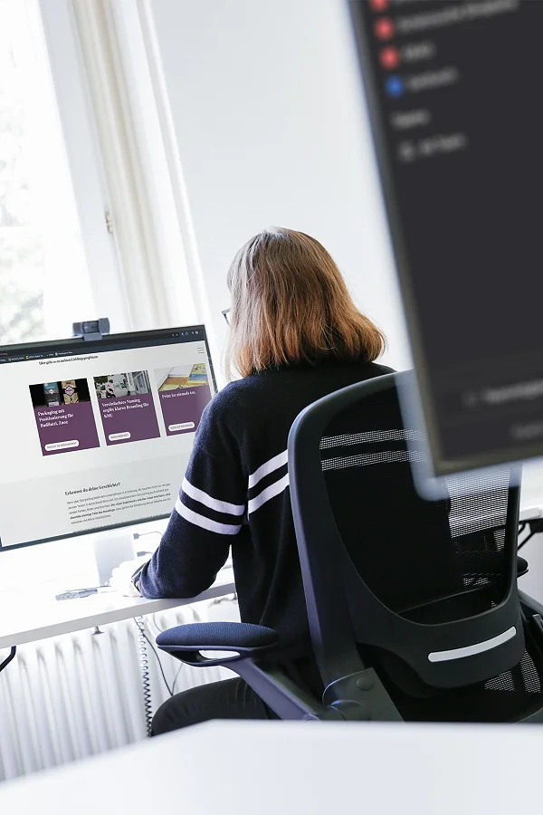 Mitarbeiterin am Schreibtisch vor zwei Bildschirmen am Fenster