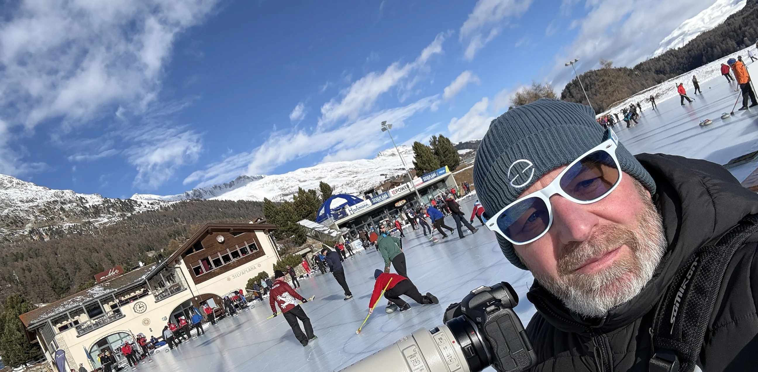 Roger mit Teleobjektiv auf dem Stativ am Curling-Rink in Silvaplana, dahinter das Spielfeld und die Bergkulisse