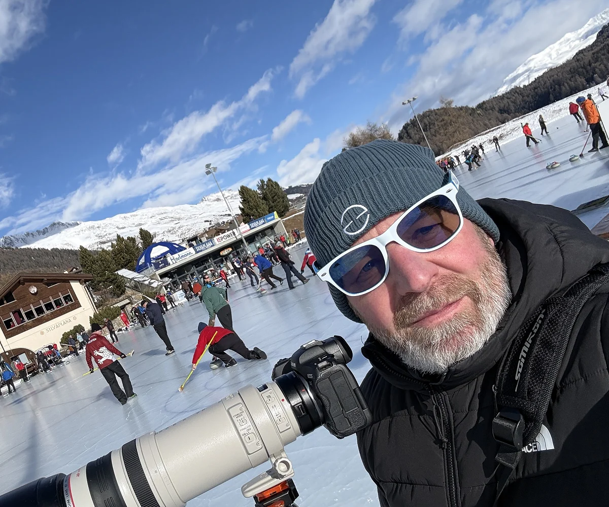 Roger mit Teleobjektiv auf dem Stativ am Curling-Rink in Silvaplana, dahinter das Spielfeld und die Bergkulisse