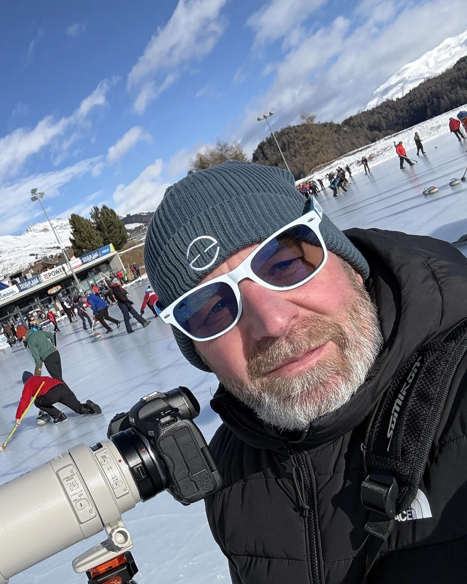 Roger mit Teleobjektiv auf dem Stativ am Curling-Rink in Silvaplana, dahinter das Spielfeld und die Bergkulisse