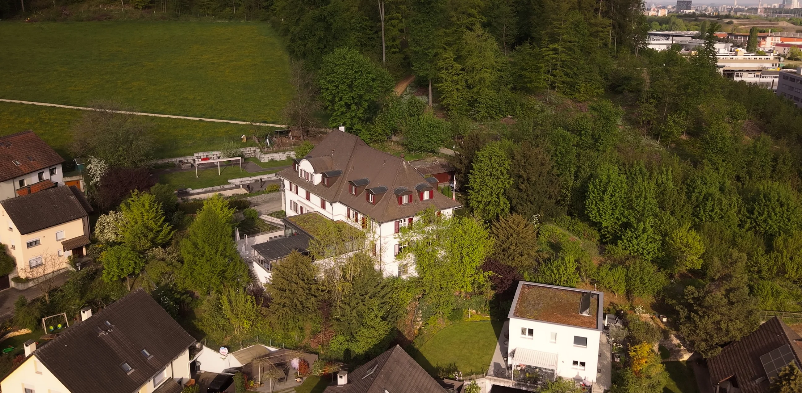 Drohnenaufnahme eines Adulta-Standorts: Villa mit Terrasse am bewaldeten Hang, im Dunst dahinter die Stadt