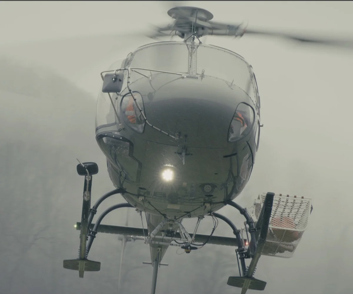 Helikopter im Schwebeflug von vorn, mit Lastseil und Transportkorb, dahinter neblige Bewaldung