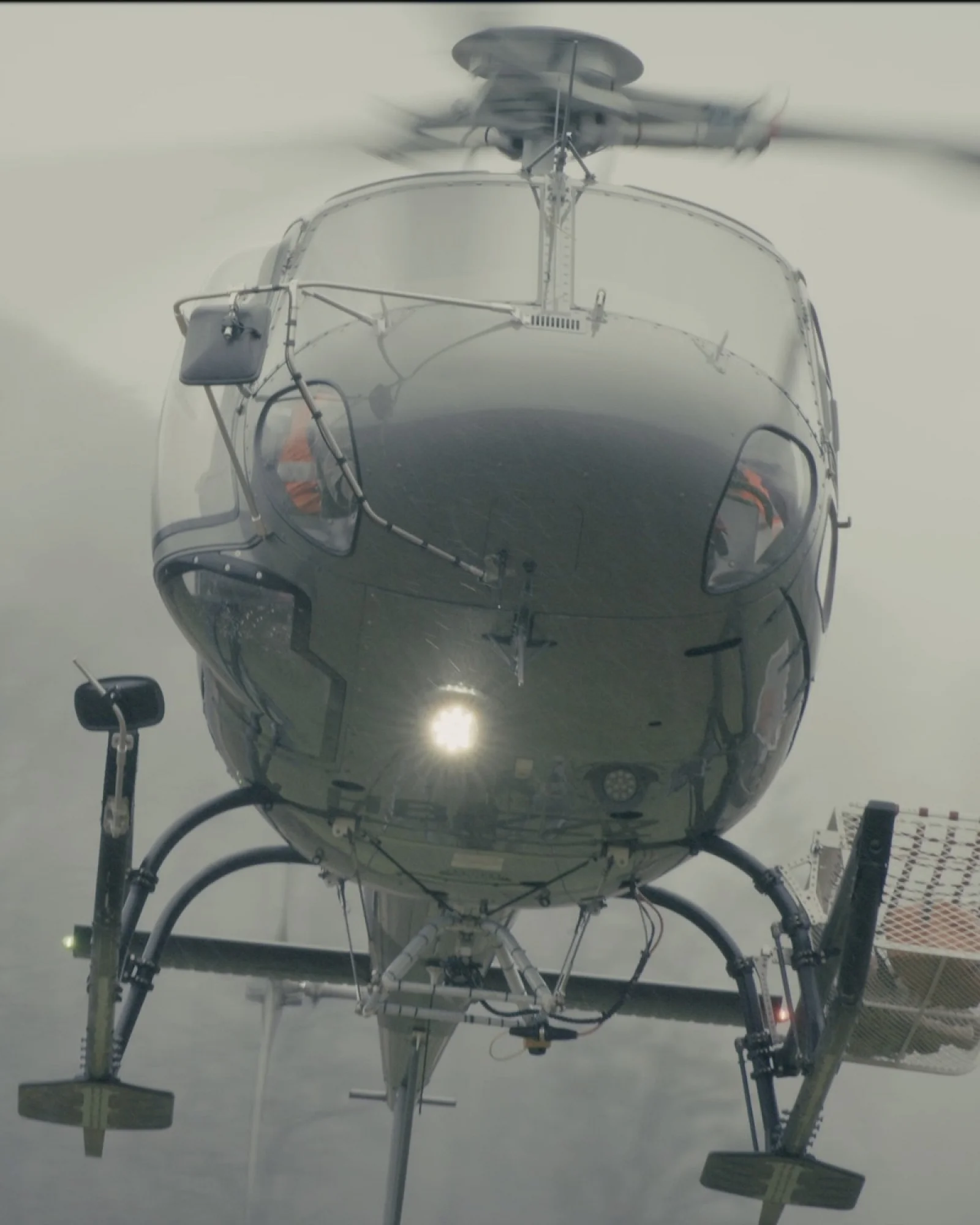 Helikopter im Schwebeflug von vorn, mit Lastseil und Transportkorb, dahinter neblige Bewaldung