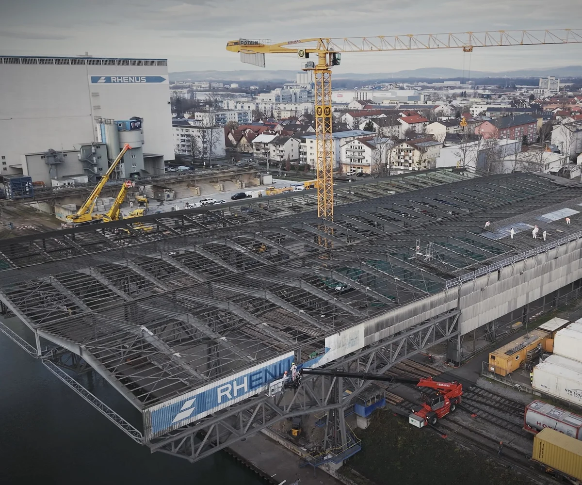 Drohnenaufnahme des Rückbaus: Hallendach bis auf das Stahlskelett abgetragen, Arbeiter in Schutzanzügen, dahinter Rhenus-Silo und Hafen