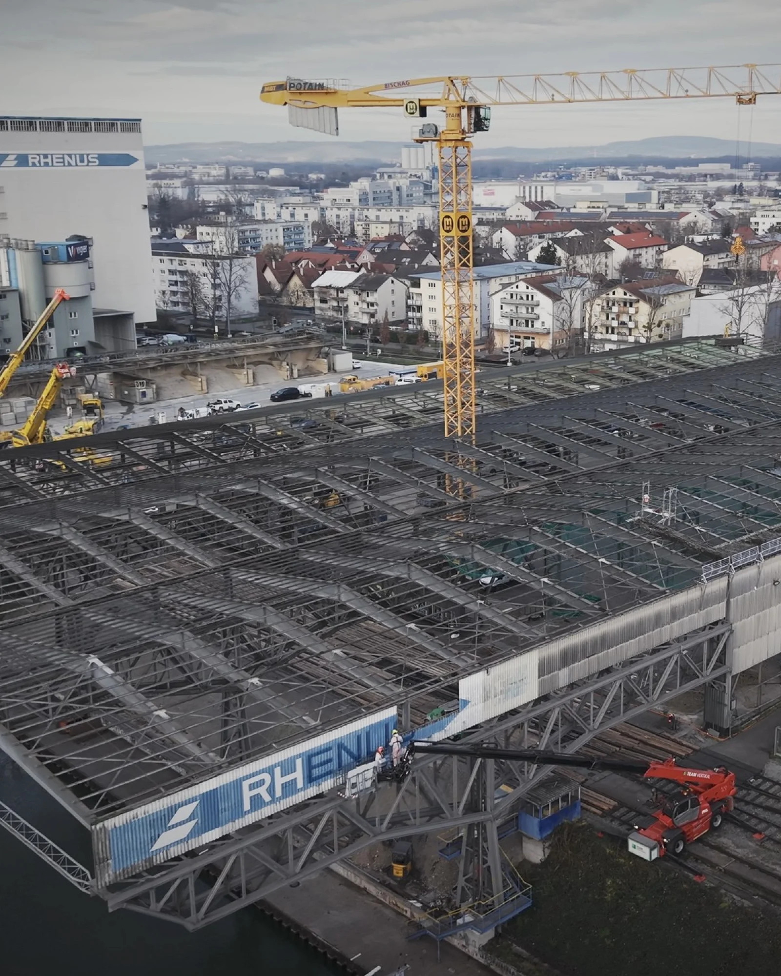Drohnenaufnahme des Rückbaus: Hallendach bis auf das Stahlskelett abgetragen, Arbeiter in Schutzanzügen, dahinter Rhenus-Silo und Hafen